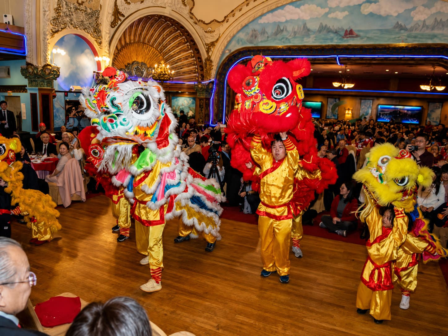 Lion dance troupes in red, yellow and white costumes performing between banquet tables in the packed theatre hall, guests watching from their seats.