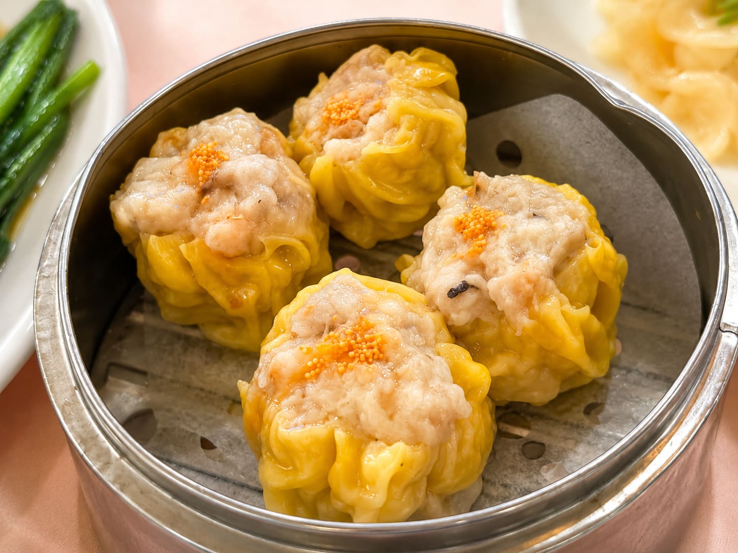 Five open-topped siu mai dumplings in a steel steamer basket, each finished with a dot of orange roe.